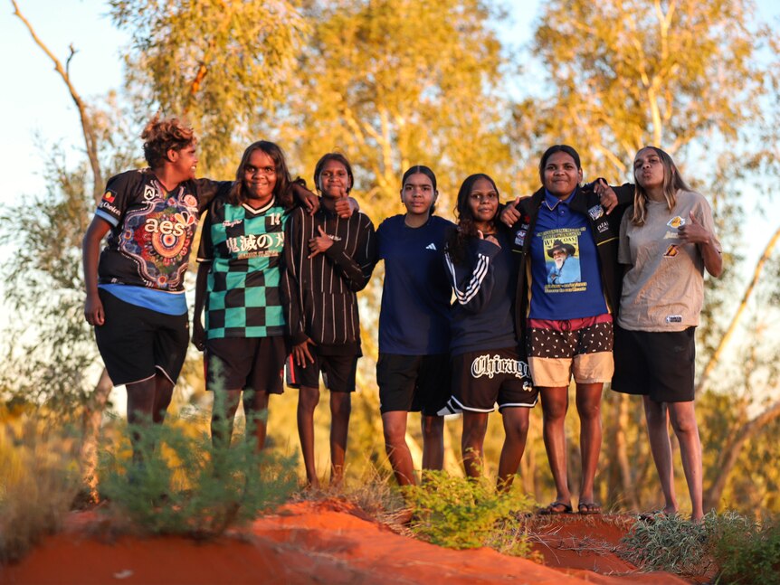 A group of young Aboriginal women wearing casual clothes standing barefoot on red sun dunes in late afternoon sunlight