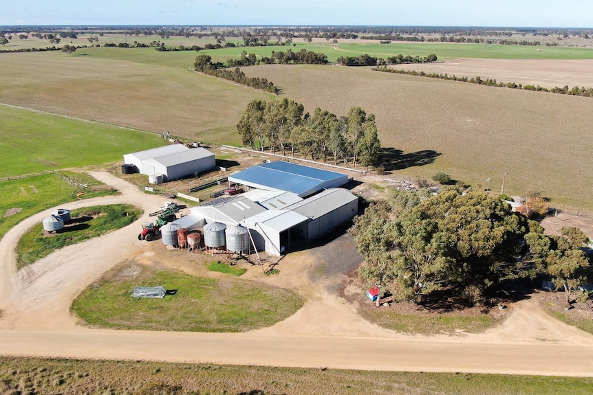 A drone shot show a eight large sheds, a series of water tanks and a house surrounded by large trees on a sprawling farm.