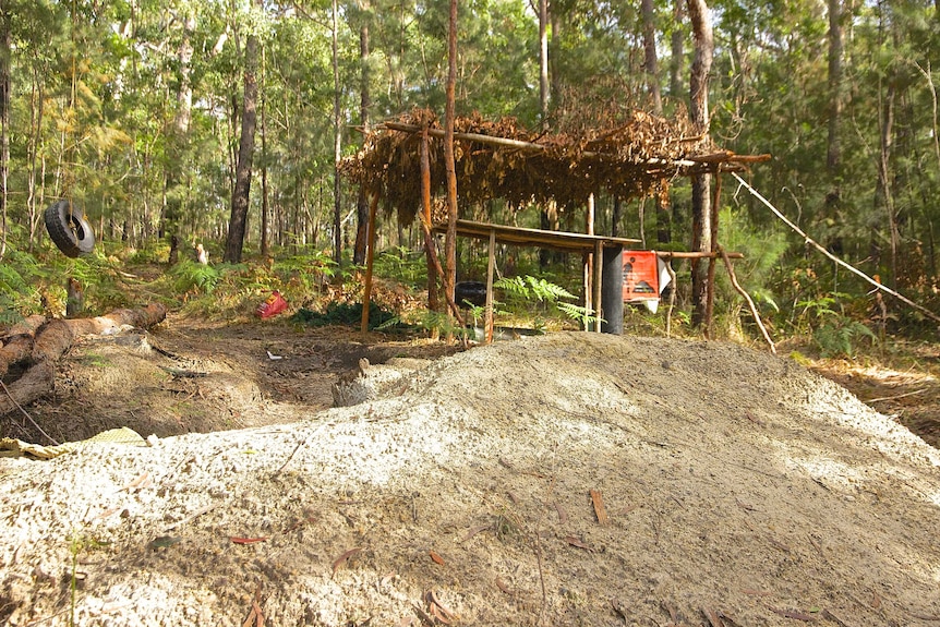 In lush bushland, there is earthworks, a make-shift pergola, construction signs, a BBQ a role swing and rubbish scattered.