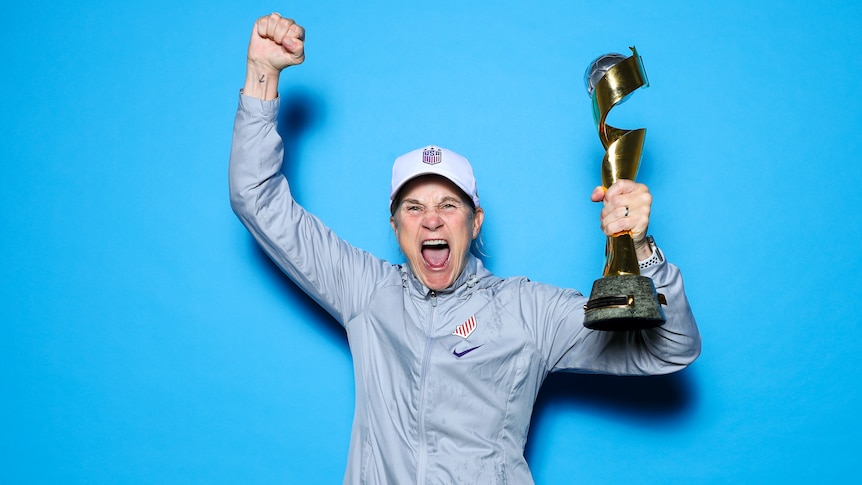 A woman wearing a grey jacket and white hat holds a trophy and yells