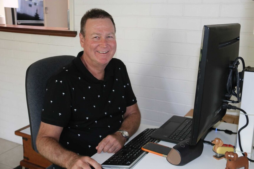 A middle-aged man wearing a black polo shirt sits at the computer in his loungeroom smiling at the camera.