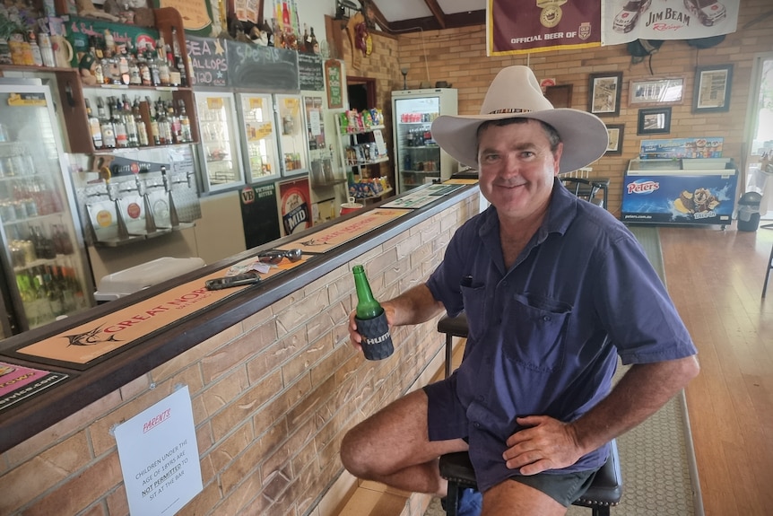 Wide shot of a man in a workshirt and hat sitting at a pub bar holding a beer bottle