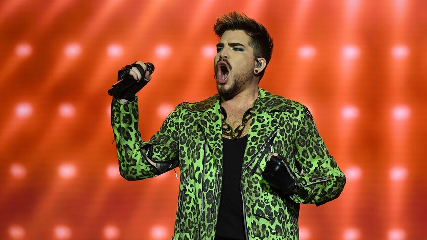 Adam Lambert in a green leopard-skin jacket sings with an orange background.