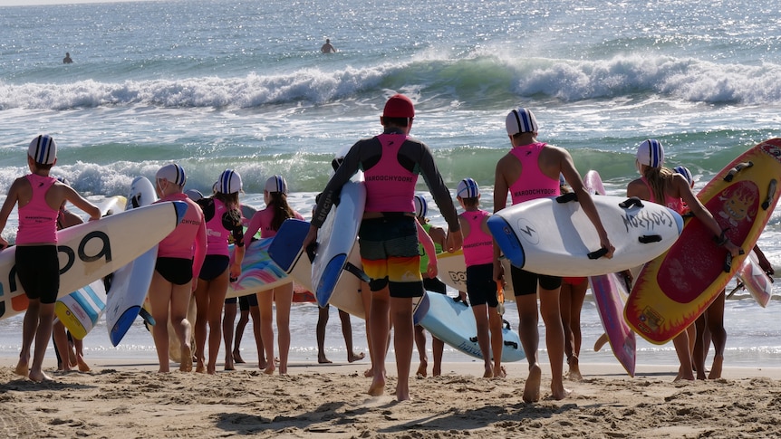 Dozens of children in fluoro vests walk walk towards the ocean, carrying surfboards.