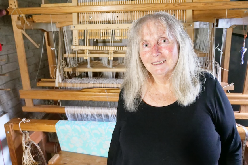 A huge wooden loom with many white threads fills a room behind a lady in her 60s with long grey hair.