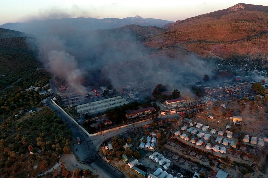 At sunrise you view a smoking migrant camp nestled between a mountain range.