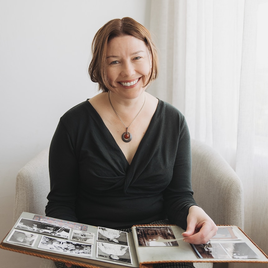 author andra putnis sits on a white chair holding a book of family photos on her lap. 