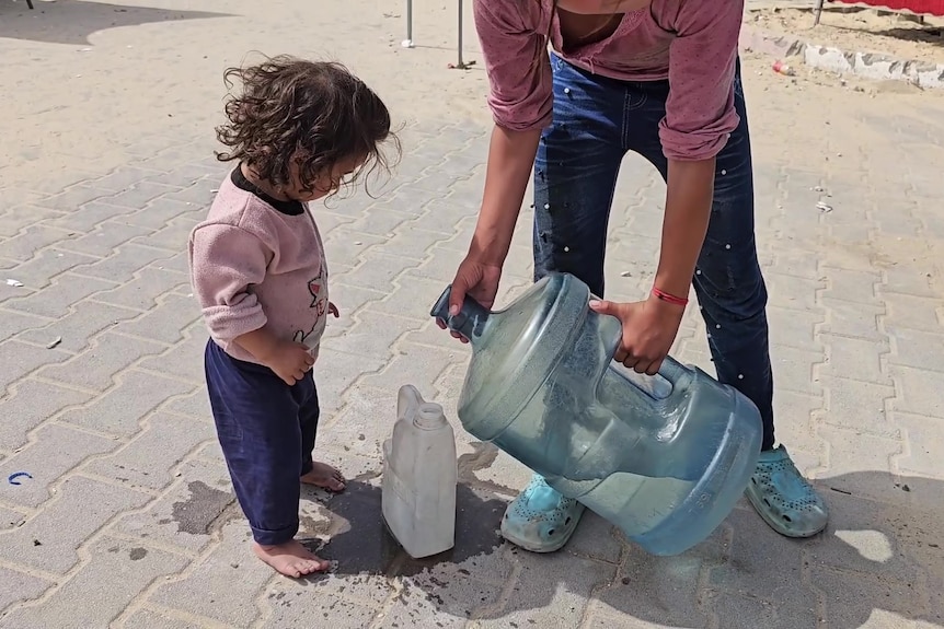 A small girl stands looking at a small bottle being filled with dirty water by another girl