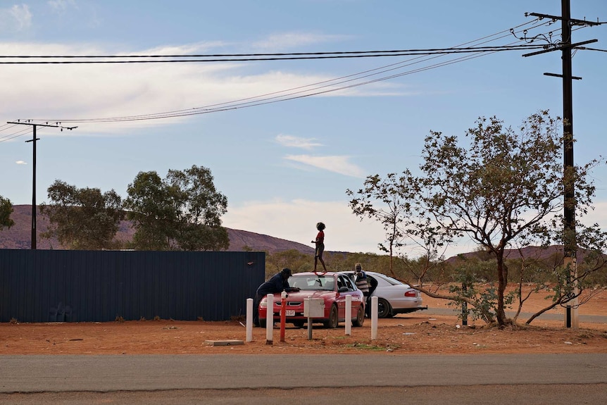 A child climbs on a red car in the middle distance with a mountain behind and powerlines above.