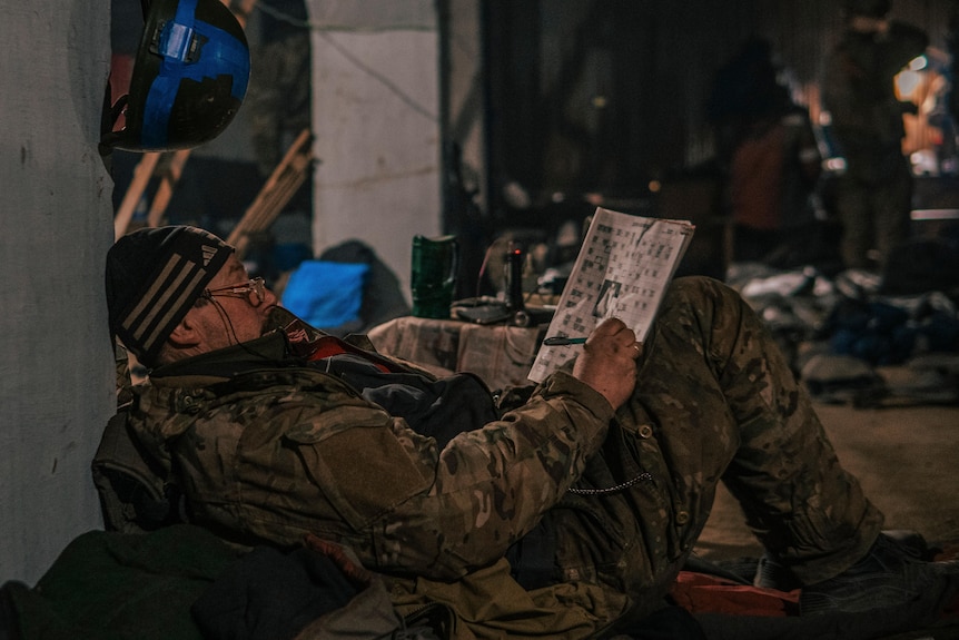 Man on the ground leans against wall and does crossword puzzles.