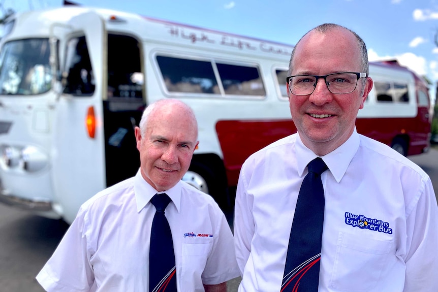 Two middle aged white men stand in front of a red and white old fashioned bus.