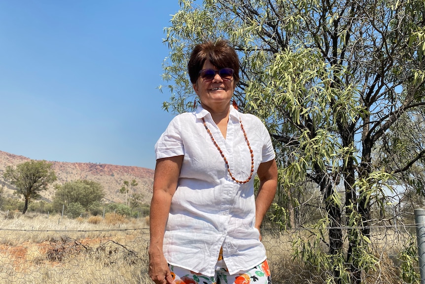 The camera looks up at a woman wearing a white shirt, Ininti seed beads and sunglasses in front of a fruitless quandong tree 