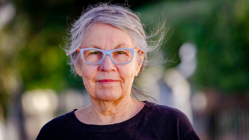 A woman with bight blue and orange glasses a loosely tied grey hair poses with a determined look