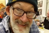 An older man in a beanie and spectacles, smiling warmly.
