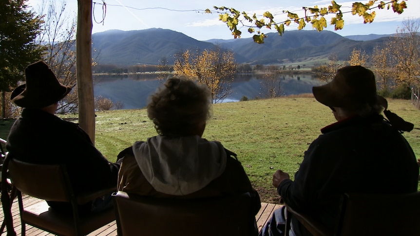 Two men and a woman (all silhouetted) sit and look at the view of a lake.