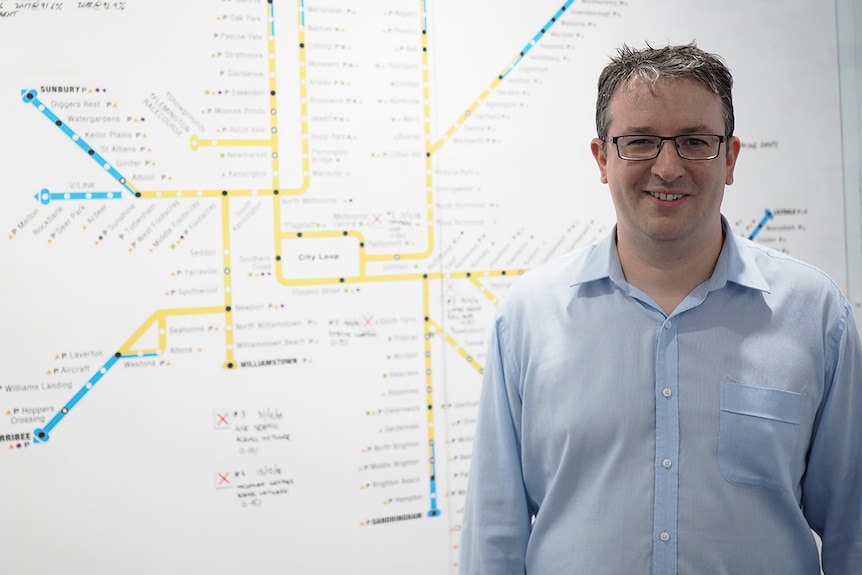 A man in a blue business shirt and glasses stands in front of a large map of Melbourne's train network.
