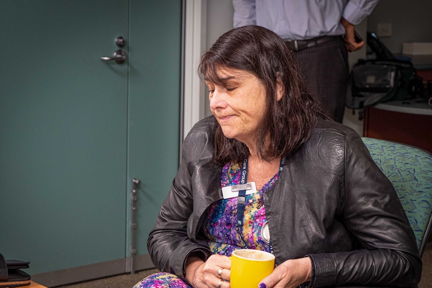 Catherine Morley holding a cup of coffee while sitting in a meeting, she looks down at her notes with a slight grimace.