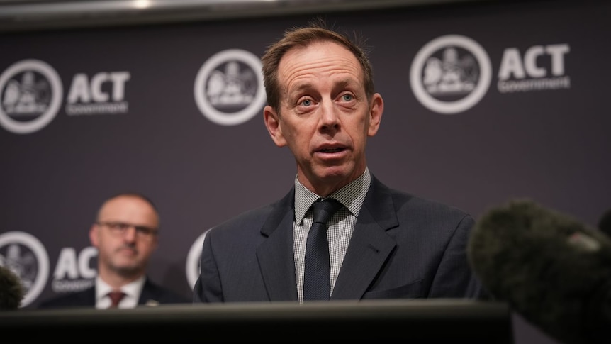 ACT Attorney-General Shane Rattenbury fronting media in a suit at a lectern in front of an ACT government backdrop.