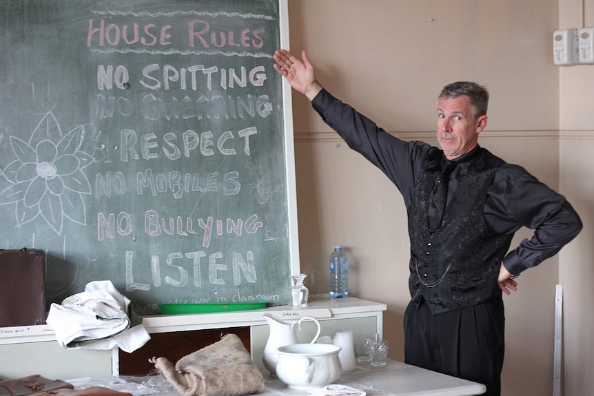 A man stands beside an old school room blackboard and points to the house rules written there