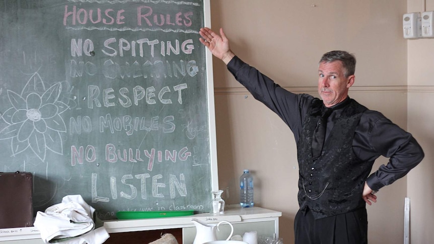 A man stands beside an old school room blackboard and points to the house rules written there