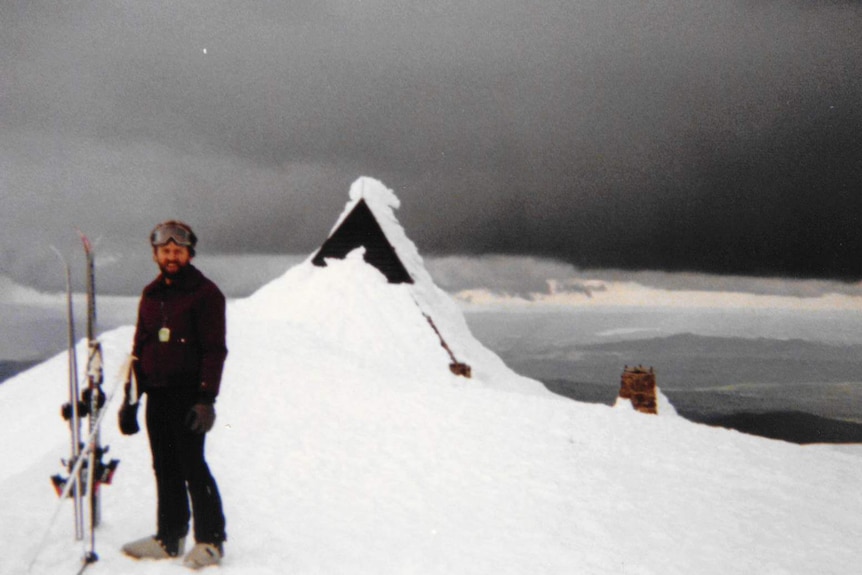 A younger Conrad Whitlock stands in ski gear on the snow, with his skis standing up next to him.