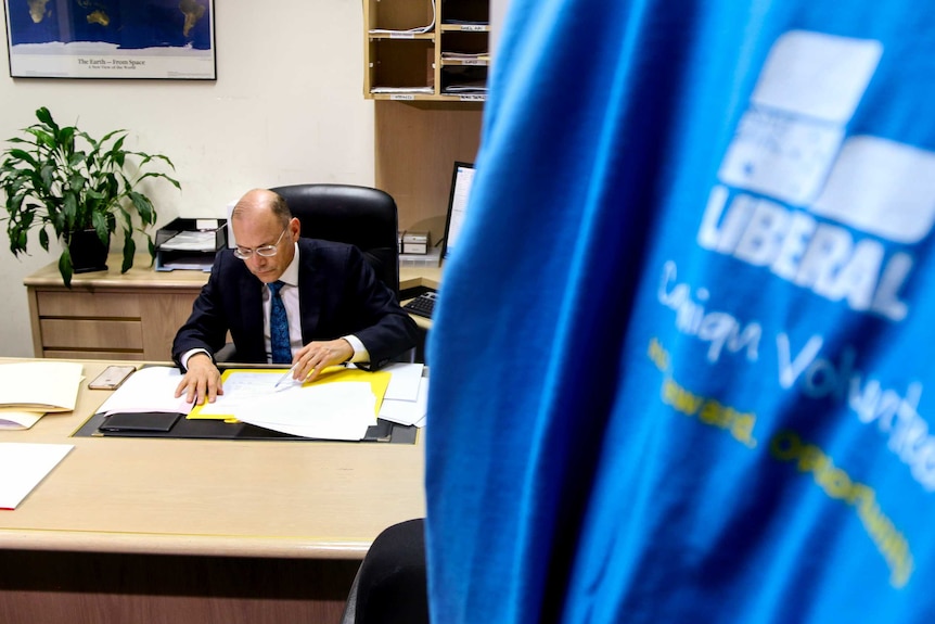 Man wearing suit and glasses sits at desk in office, looking at documents, with blue Liberal Party t-shirt hanging in foreground