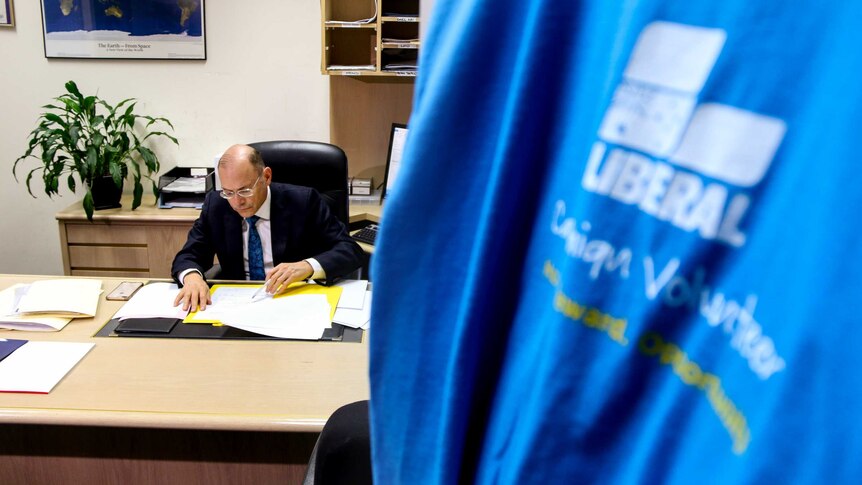 Man wearing suit and glasses sits at desk in office, looking at documents, with blue Liberal Party t-shirt hanging in foreground