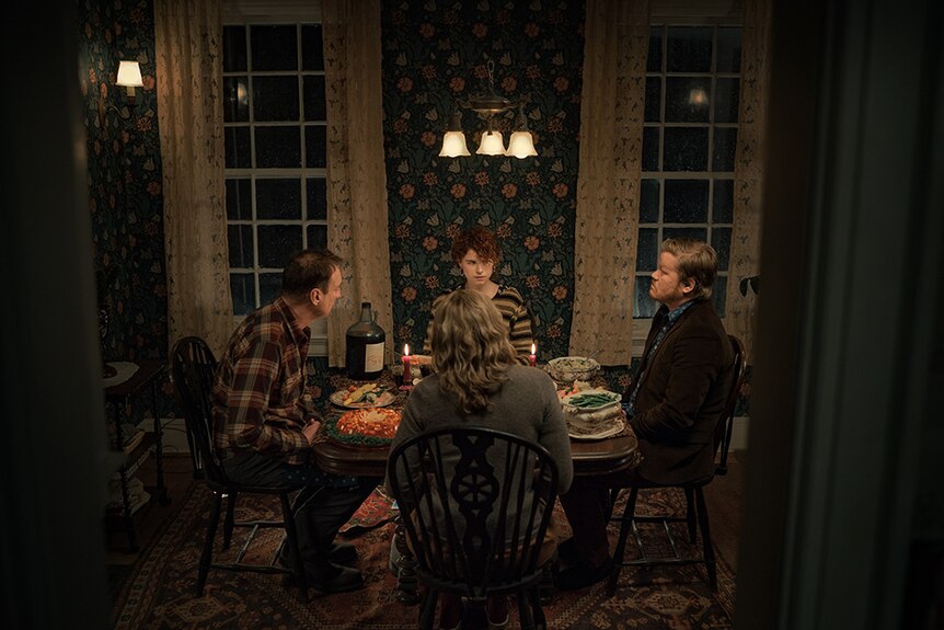 An older woman and man and younger woman and man sit at square dining table in floral wallpapered interior room at night time.
