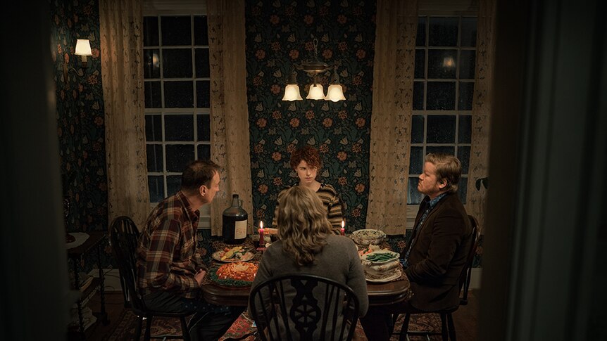 An older woman and man and younger woman and man sit at square dining table in floral wallpapered interior room at night time.