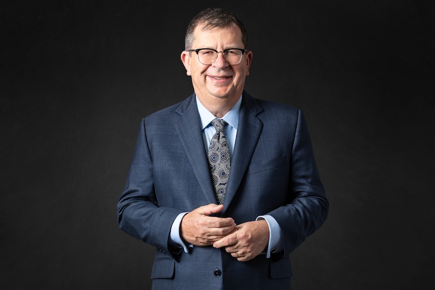 A photo of a middle-aged man in a dark blue suit and tie, wearing glasses and smiling