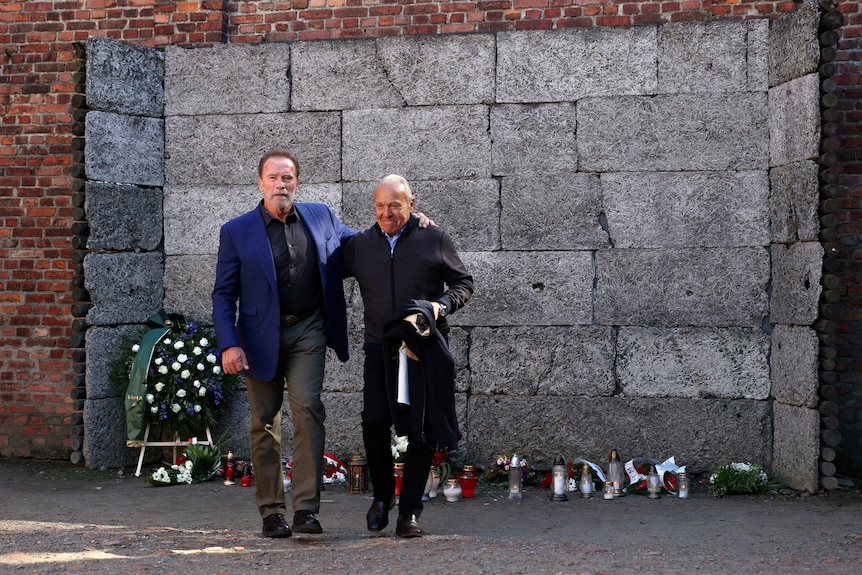 Arnold Schwarzenegger, left, and another man walk in front of wall of death in Auschwitz.