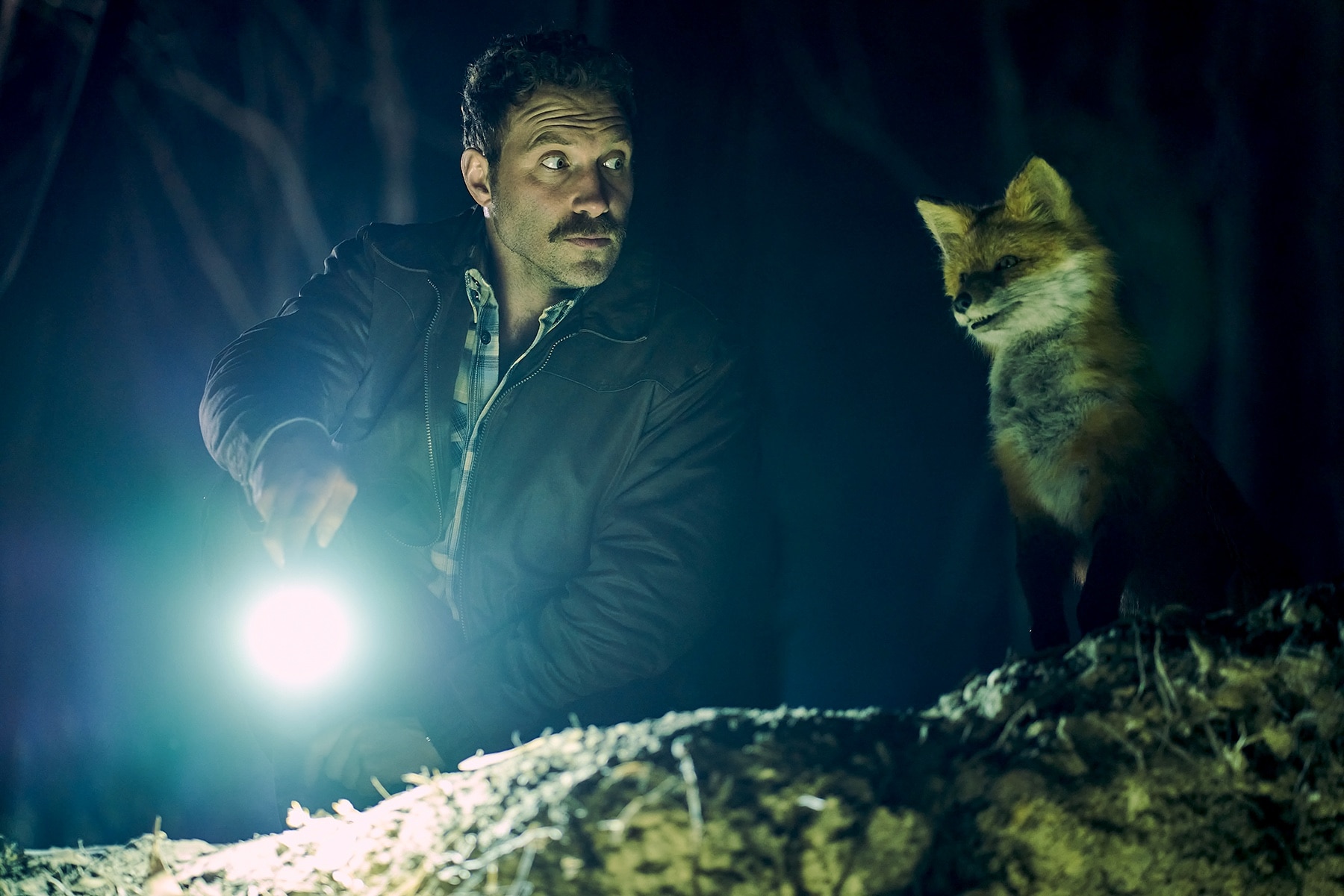 Jai Courtney stands in the dark with a torch next to an animated fox