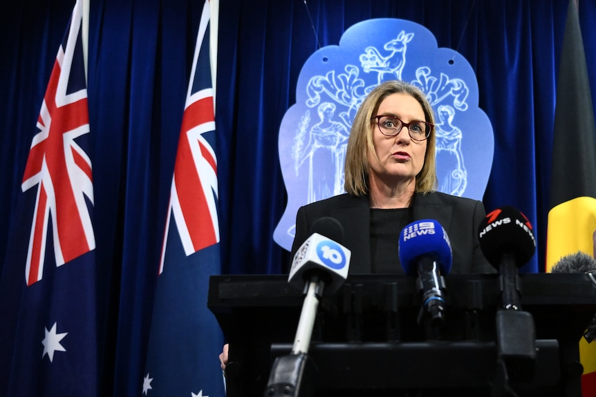 Jacinta Allan stands at a lectern with microphones in front of her. Behind her is the Australian flag