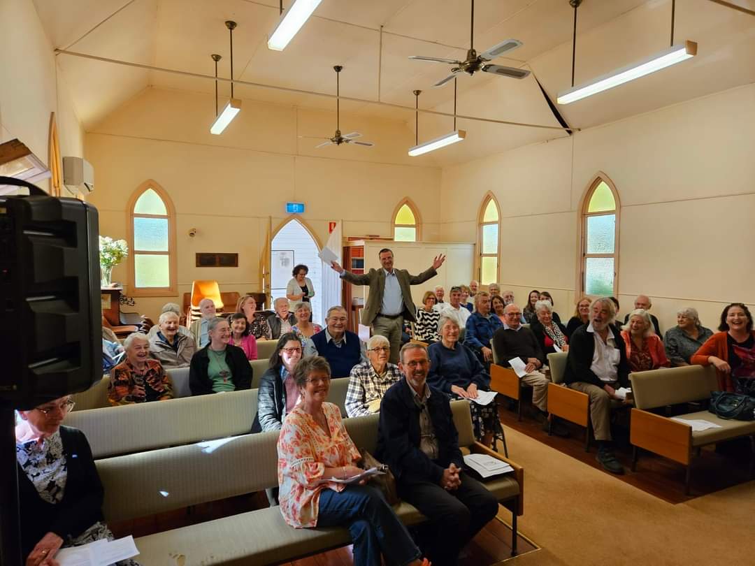 Interior of church, conical stained glass windows, fans, people sit on padded pews, a man stands in centre, arks akimbo.