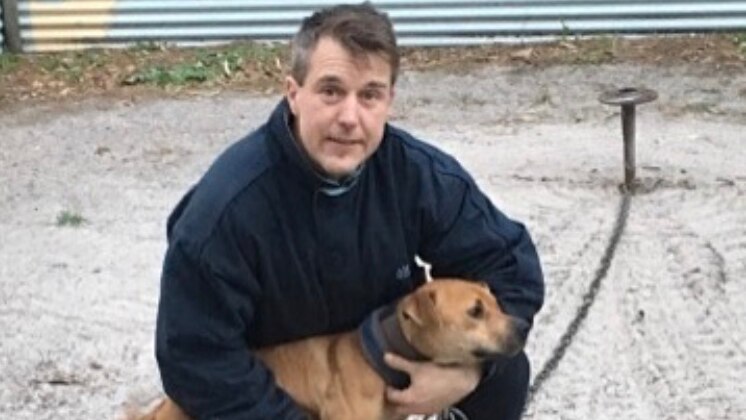A man kneels down holding a dog wearing a chain.