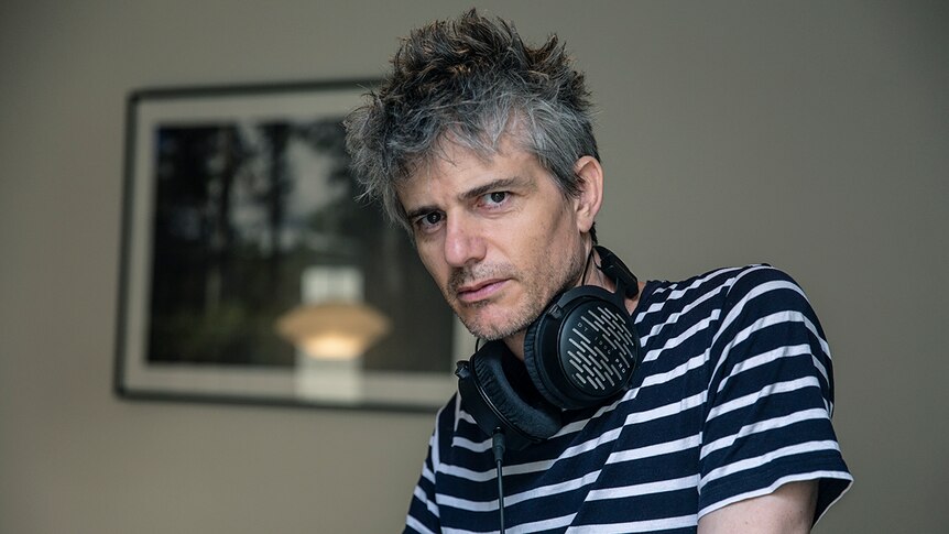 A grey-haired middle-aged man with headphones around his neck gazes moodily into the camera