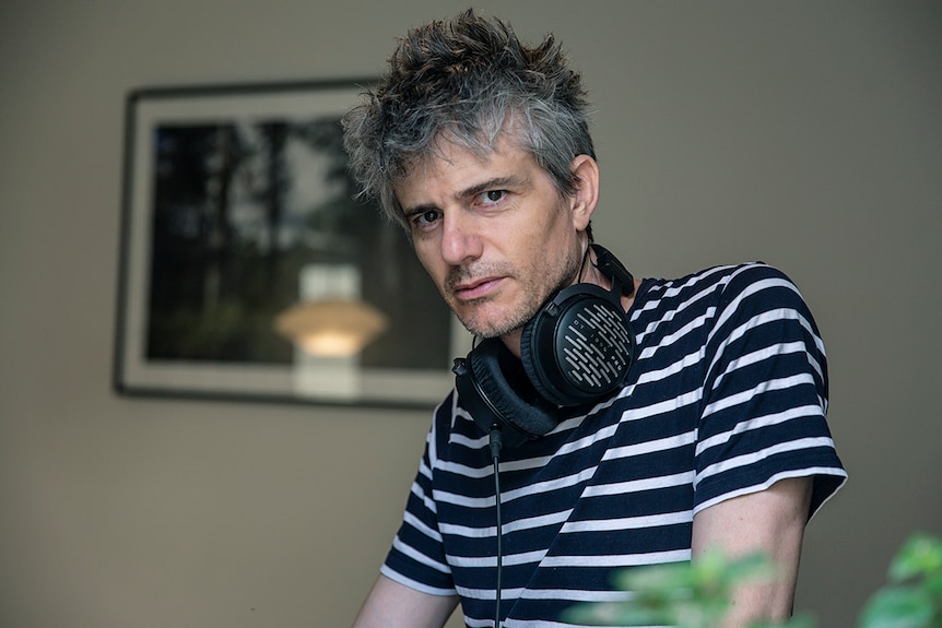 A grey-haired middle-aged man with headphones around his neck gazes moodily into the camera