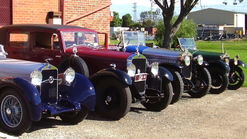 A group of old cars, one blue, one red, one silver and one green are parked in a line by a brick wall.