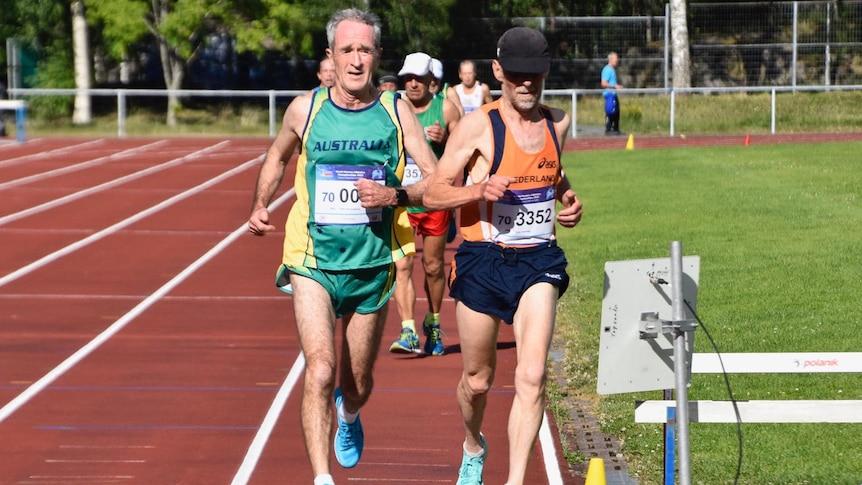Two men running on the inside lane of an athletics track, one wearing an orange and black shirt, the other a green and gold