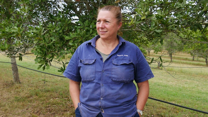 A middle aged woman stands proud, hands in pockets in a macadamia orchard. She dons an old work shirt and pants.