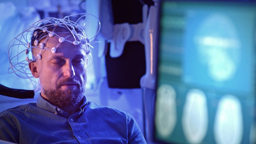 A man with his eyes closed and a headset of electrodes on his head. In the foreground out of focus is a screen with brain images
