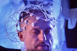 A man with his eyes closed and a headset of electrodes on his head. In the foreground out of focus is a screen with brain images