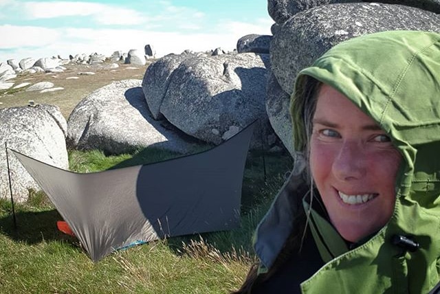 Smiling woman, pictured in cold weather gear, takes a selfie with large boulders and a small shelter in the background.