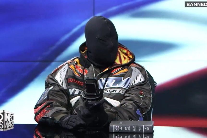 Kanye West sits before a microphone wearing a black mask and racing jacket, an American flag graphic behind him.