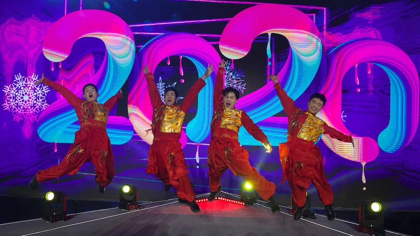 Four men jump in front of a large pink sign reading 2020.