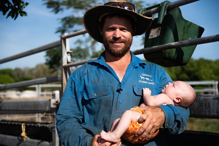 A smiling, bearded man in a work shirt and hat holds a tiny baby.