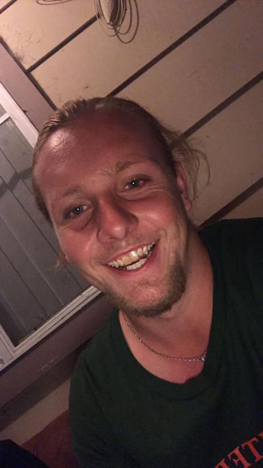 A close-up of a smiling young man with long, shaggy hair, wearing a black T-shirt