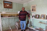 An elderly man stands in the corner of an old room with pressed metal walls and a low ceiling, surrounded by various objects.