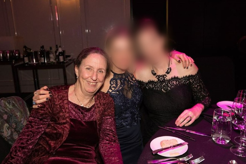 A woman in a dress poses at a dinner table alongside two other women with blurred faces. 
