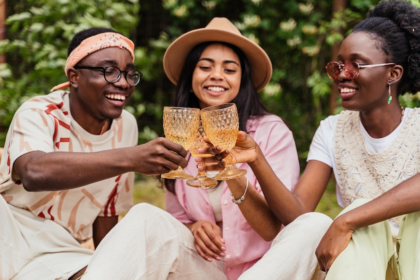Three people sit in the park smiling, wearing linen, and cheers their three glasses together